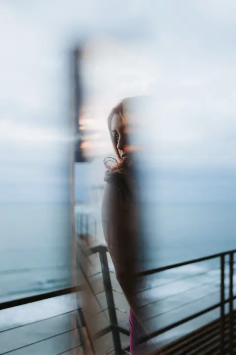 Long Truong A woman photographed through glass. The image is blurred around the woman but she can be partly seen through the glass. She is looking over her shoulder and appears to be standing on a promenade beside the sea.