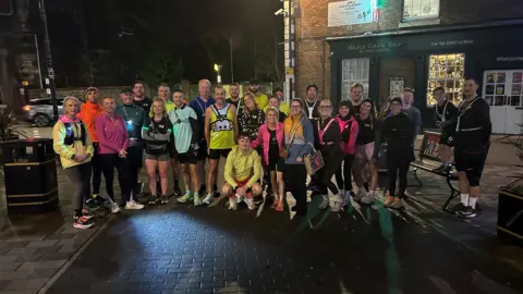 A gathering of people in multi-coloured running gear in a paved town square with a shop behind them
