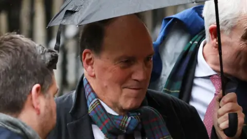 Sir Simon Hughes pictured holding an umbrella over his head outside the High Court in London. He wears a dark coat and tartan scarf.