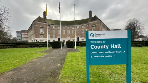 The outside of County Hall in Taunton. There is a pavement and a green lawn in the foreground along with a sign saying "Welcome to County Hall." There is a large building in the background along with three flag poles.