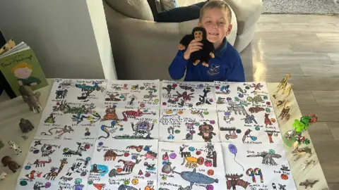 Samantha Evans-Browning A little boy in a blue school uniform holds a cuddly monkey toy and in front of him are A3 pieces of paper joined together with animals drawn on them