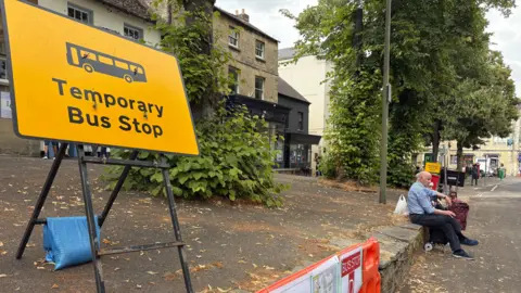 A big yellow temporary bus stop sign on a hill in Witney. Trees line the street and an elderly couple sit on the wall beside the sign as they wait for their bus