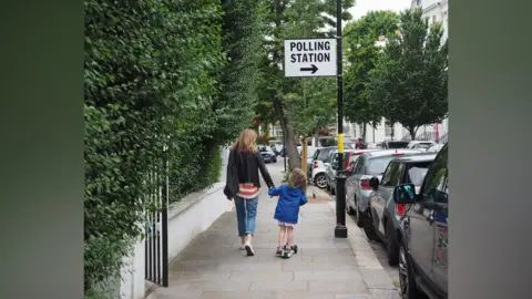 A woman and a young child walking down a tree-lined suburban street hand in hand. Attached to a lamppost is a sign that says 'polling station'.