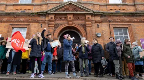 People have gathered outside the library. Some have banners and a woman has a loudspeaker. The protesters are wearing outdoor coats and shoes. The decorative entrance to the library is behind them.
