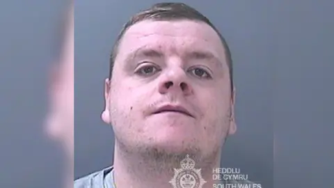 Police custody still of Ashley Davies. He is looking at the camera and there is a South Wales Police logo in the bottom right hand corner