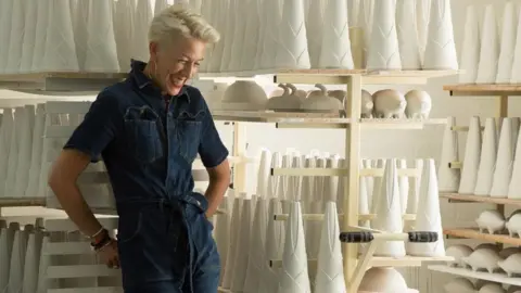 A woman wearing dark blue denim overalls leans against a shelf and smiles while looking down. Behind her are shelves filled with white ceramic pieces.