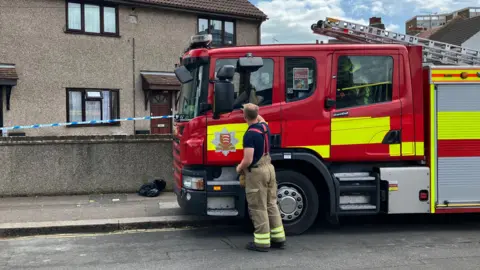 Henry Godfrey-Evans/BBC A red fire engine parked outside a house. There is also a police cordon in place.