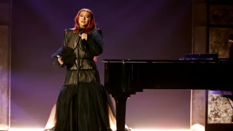 Getty Images Woman in black dress by a piano. She has red hair and holds a microphone.