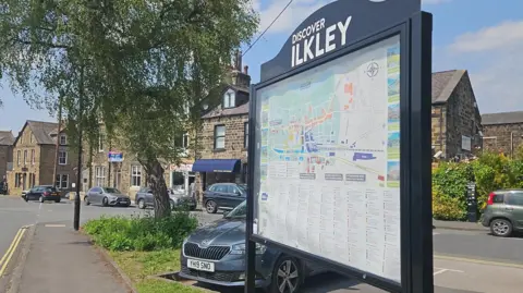 Aisha Iqbal/BBC A big street information sign in a traditional English style market town. The sign says 'Discover Ilkley' at the top and has a map and a long list of tourist information.