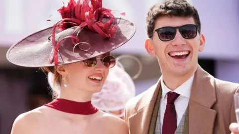 PA Media A woman is wearing a dark red dress, fascinator and sunglasses. She is stood with a man who is wearing a tweed suit, beige coat and sunglasses. Both are smiling