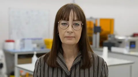A woman with brown hair is wearing a glasses as well as a brown and white striped shirt. She is standing in what appears to be a factory. The room is brightly lit, and there are work benches and equipment behind the woman.