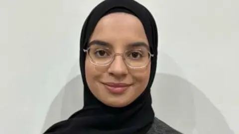 University of Cumbria Umairah Malik stands in front of a white background. She wears clear-framed glasses and a black headscarf.