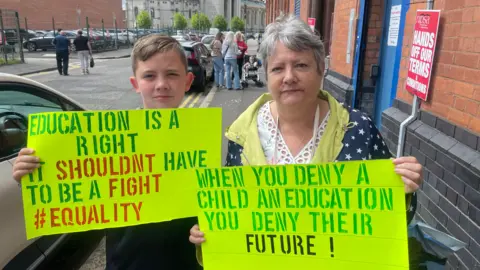 BBC Ethan and his grandmother Isobel Woodhead holding yellow placards outside the Education Authority's HQ in Belfast. Ethan has short, blonde hair which is swept across his forehead. His placard reads: "Education is a right, it shouldn't have to be a fight. #Equality. Ms Woodhead has short, grey hair and is wearing a navy rain mac decorated with white stars over a white top.