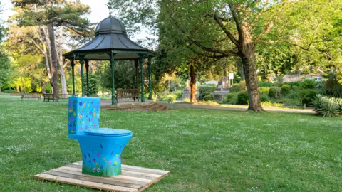 Sienna Anderson A blue toilet patterned with colourful flowers. A bandstand is in the background.