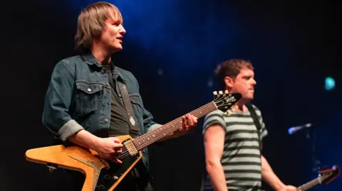 Getty Images A man with brown hair and a denim jacket holding a brown guitar with a man with brown hair and a striped grey tshirt behind him