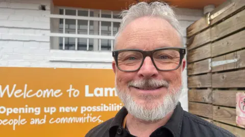 A bearded man with grey hair and glasses stands in front of a council sign