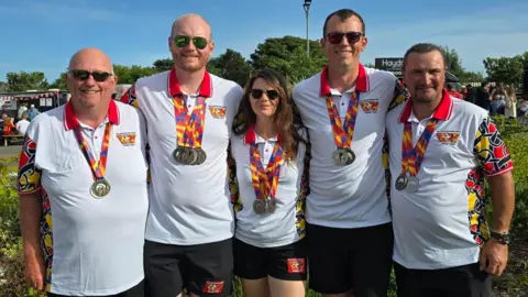 Isle of Man Archery Team Five people in white T-shirts stand with their arms around each other. They each have medals on colourful ribbons around their necks.