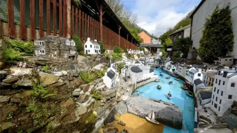 Some of Polperro Model Village, featuring various white-fronted model buildings along two banks of a narrow river, with the river having a harbour wall and a number of boats on it