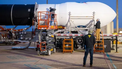 Firefly Aerospace/Trevor Mahlmann Engineers work on the nose of a large rocket that is lying horizontal on a support structure.