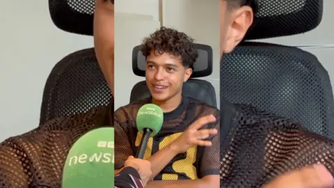 A young man with short curly hair. He's smiling and has a green microphone in front of him.