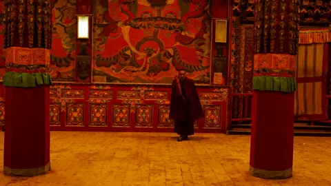 Xiqing Wang/ BBC A monk inside a monastery whose walls are covered in murals in rich reds.
