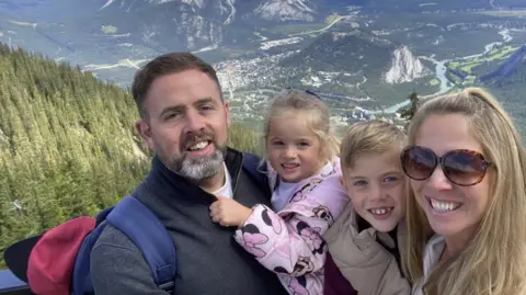 Family handout A family stand in front of a dramatic view down a steep valley. Trees can be seen on the left, with a river snaking through the valley. A man and woman smile at the camera with two young children. 