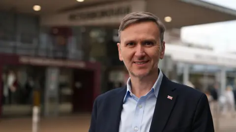 MARTIN GILES/BBC Andrew Pakes is smiling and looking at the camera. He is wearing a light blue shirt and navy blazer with a Union flag pin. Behind him is the entrance to Peterborough's railway station.