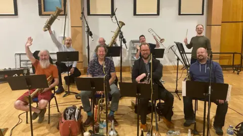 Jamie MacDougall Eight people who make up a brass ensemble. They are sitting at music stands and holding their instruments in front of them or raised in the air. One of them is a conductor with his baton