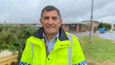 Police and Crime Commissioner for Wiltshire and Swindon, Philip Wilkinson, wearing a hi-vis jacket with police checkerboard on. He is smiling into the camera and is stood beside a road.