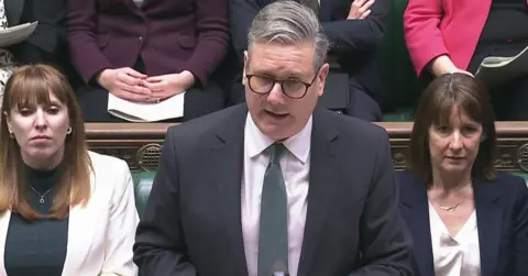 The Prime Minister at the Dispatch Box during Prime Minister's Questions, Wednesday March 5th 2025. He is wearing a gray suit. Sir Keir Starmer is flanked by Angela Rayner and Rachel Reeve. 