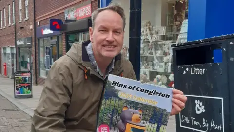 A man in a town centre, poses next to a bin with a calendar. He is wearing a brown coat over a checked shirt. The calendar is titled 'Bins of Congleton 2025' and its main image is of a bin which resembles a bear.