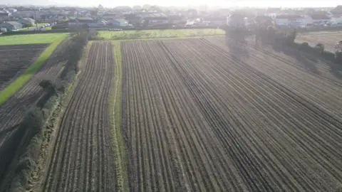 BBC An aerial view of ploughed farmland with houses in the far distance