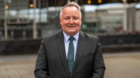 Darren Millar MS is looking towards the camera wearing a grey suit with a sky blue shirt as well as a green spotted tie. 
