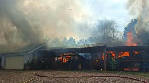 Shropshire Fire and Rescue Service A farm outbuilding with agricultural vehicles is blackened and surrounded by billowing smoke. There are huge orange flames coming from it