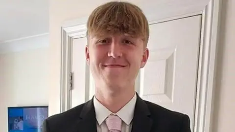 Alfie looking at the camera smiling. He is wearing a black jacket, white shirt and pink tie.