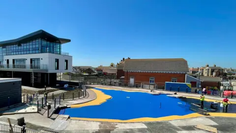 On the right hand side of the photo is a paddling pool which has only been partially filled with water. The pool floor is visible and is electric blue with a yellow border. There are workmen to the right who are finishing the maintenance work. Overlooking the paddling pool to the left hand side of the photo is the new Seaview Colonnade - a modern building dominated by a glass frontage. 