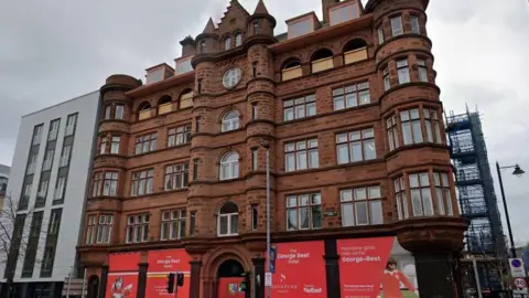 The exterior of Scottish Mutual building - a large, red brick Georgian building on the corner of Bedford Street in Belfast.  It has several tower features, dozens of windows, some curved, and is attached to a white building. There are hoardings at the front feature the footballer George Best.