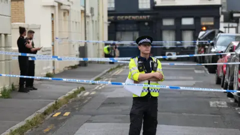 A street cordoned off by police. A police officer is stood directly behind the police tape cordon with his arms crossed. Another two officers are further into the cordon, stood on the left hand side.