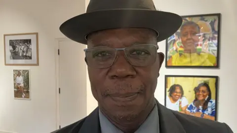 A man wearing a hat, glasses and suit standing in an art gallery, with framed photographs displayed on the walls behind him.