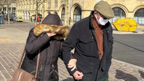 Fyona and Michael Bairstow leave Bradford Magistrates' Court. Fyona has her coat hood pulled over her face, while Michael is wearing a Covid-style face mask, dark glasses and a brown flat cap.