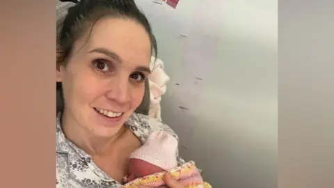 Natalie Natalie is seated indoors holding a newborn baby wrapped in a pink, yellow and white knitted blanket. The baby is wearing a light-coloured knitted hat and is nestled against the person's chest.