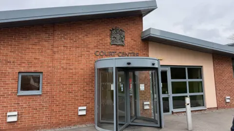 The exterior entrance of Bulford Military Court. It is a red brick building with sloped grey roofs and a revolving door. Above the door is a metal coat of arms and a sign saying "Court Centre" 