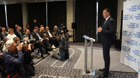 PA Media Russell Findlay stands at a podium wearing a black suit as party officials and media camera crews look on.