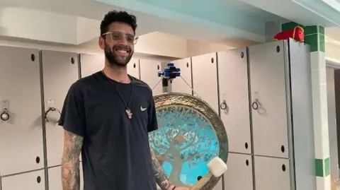 Sally Fairfax/BBC A tall man with glasses is standing in a swimming pool hall in front of the lockers. He is smiling and poised with a drumstick, ready to strike a gong, that is next to him, hanging on a stand. 
