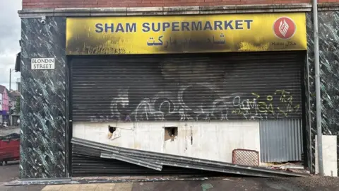PA Media The front of a shop which has been set on fire. The yellow sign has sustained smoke and fore damage and is blackened. The metal shutter has also been damaged.