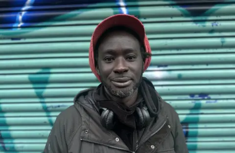 A man stands against a green door shutter. He is wearing a dark jacket and red cap