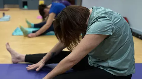Several people sit on exercise mats in a gym studio, stretching forward with legs extended. The foreground shows a person on a purple mat reaching towards their feet, with a patterned tattoo visible on one foot. Gym equipment and shelving are in the background.
