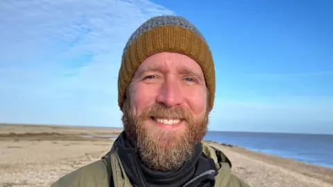 Stuart Howells/BBC Matt Wilson smiles at the camera while standing on a beach. He wears a mustard yellow and a grey beanie hat as well as a green coat with a black jumper underneath. He has a long and light-coloured beard