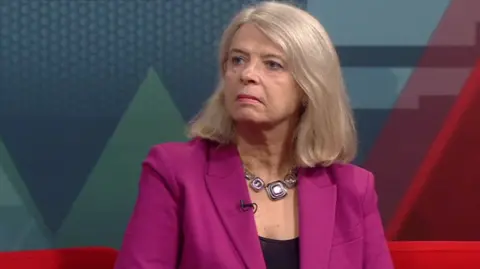 MP Dame Harriet Baldwin, a woman with shoulder-length light blonde hair, sits in the BBC Politics Midlands studio on a red sofa, while wearing a purple jacket and necklace