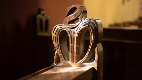 Andy Marshall The end of a church pew with a stylised s and m like a heart shape carved in the wood